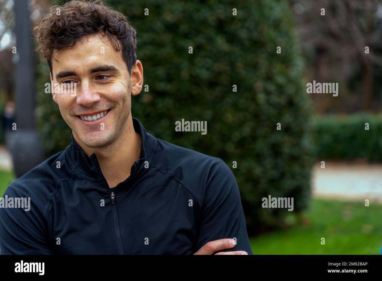 Giovane uomo positivo in camicia nera che guarda via da sogno contro sfondo sfocato del parco verde Foto Stock