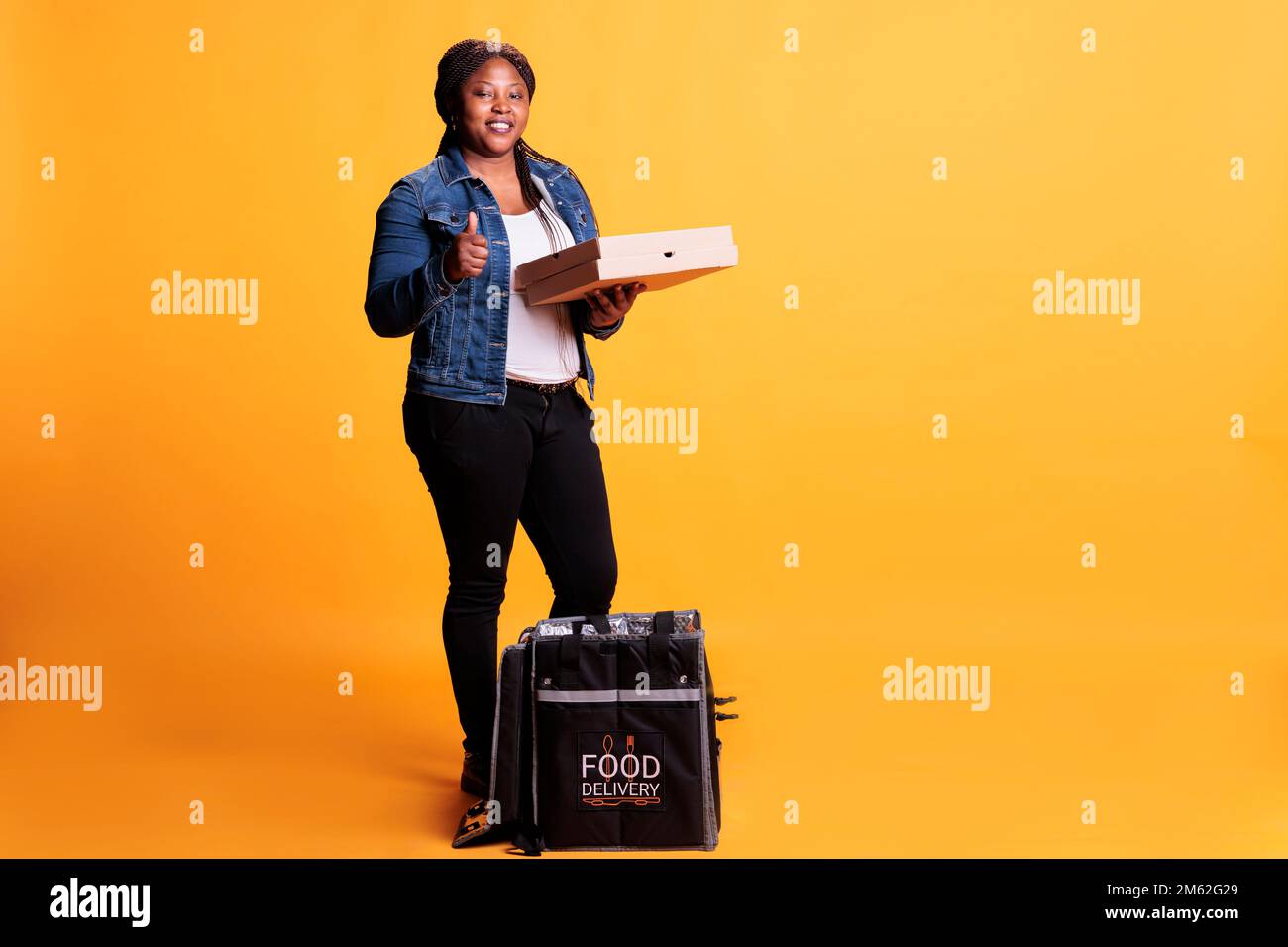 Un operatore di ristorante di successo che tiene la scatola di cartone piena di pizza mentre fa il pollice verso l'alto gesto alla macchina fotografica prima di consegna fast food ordine al cliente. Pizzeria corriere con zaino termico da asporto Foto Stock