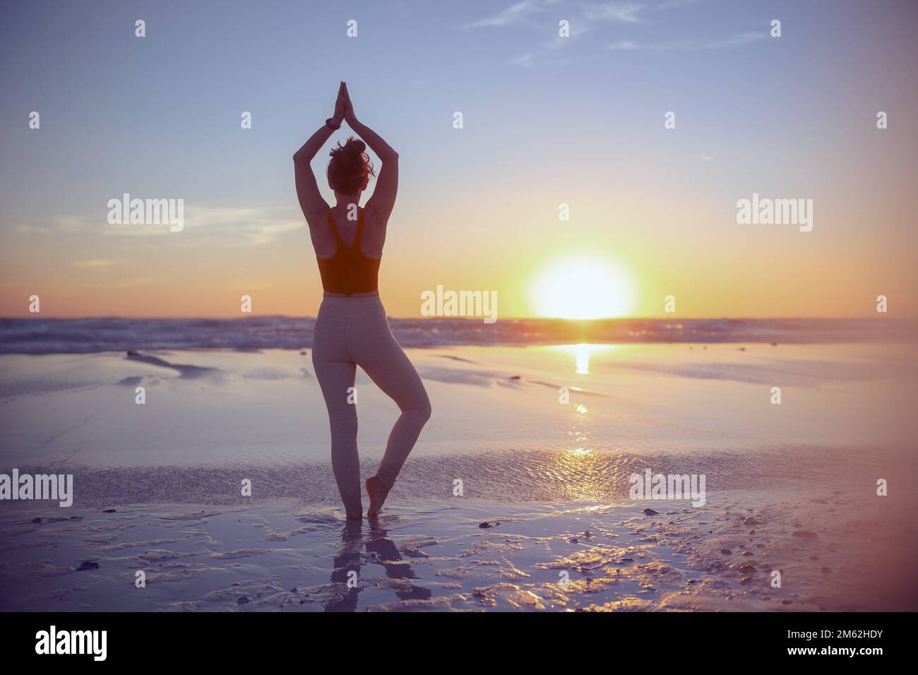 Visto da dietro palestra donna jogger in abiti fitness in spiaggia al tramonto meditando. Foto Stock