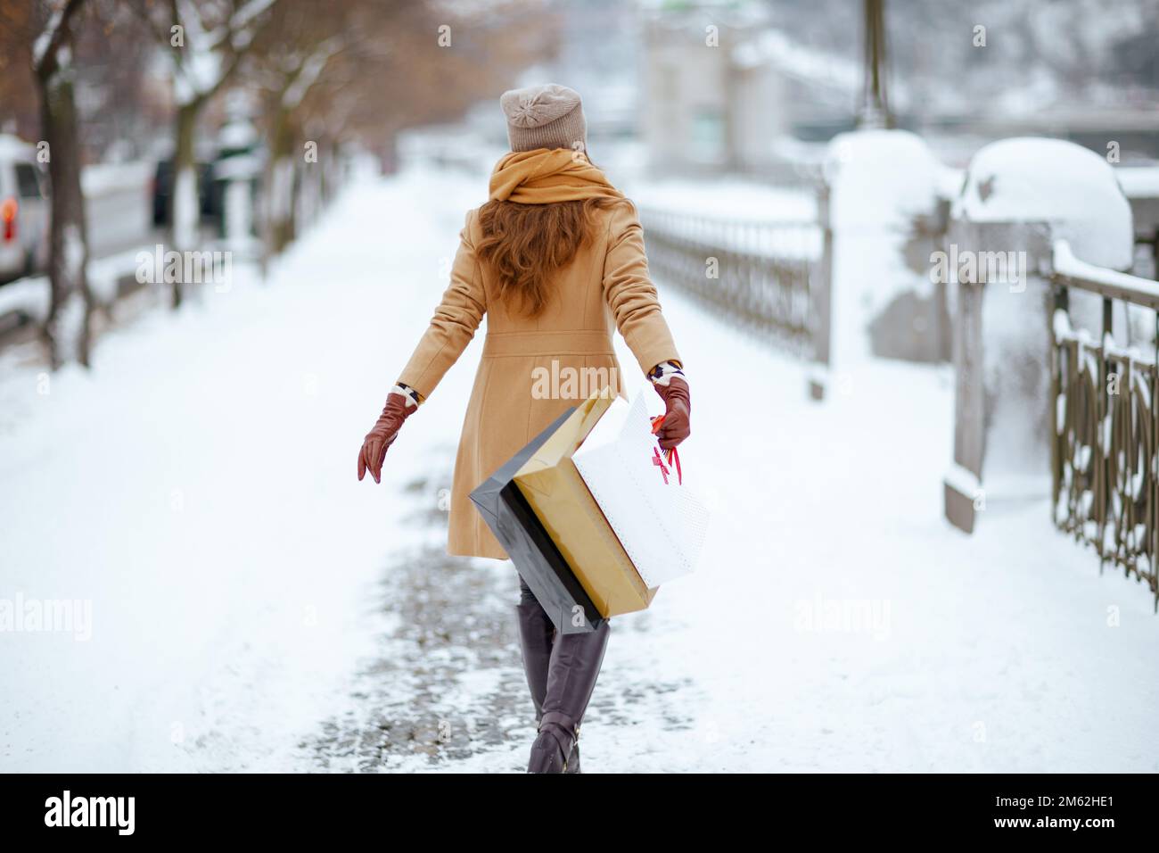 Vista da dietro donna di mezza età in cappello marrone e sciarpa in camel cappotto con guanti e borse per lo shopping fuori in città in inverno. Foto Stock