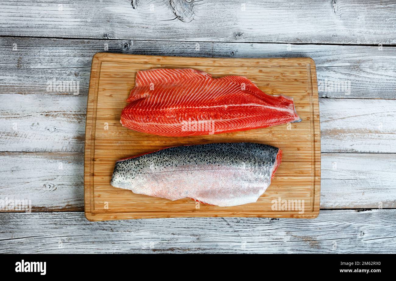 Due filetti di salmone alla trota, con la pelle rivolta verso l'alto e verso il basso, sul tagliere con fondo bianco rustico in legno Foto Stock