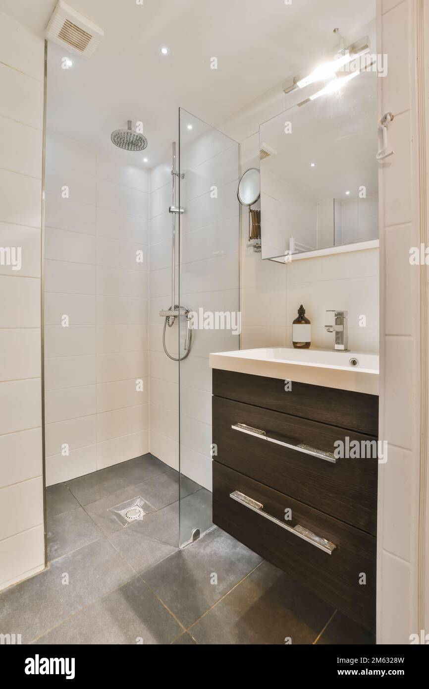 un bagno moderno con piastrelle bianche e armadietto di legno scuro sotto il lavandino si trova davanti alla porta della cabina doccia Foto Stock