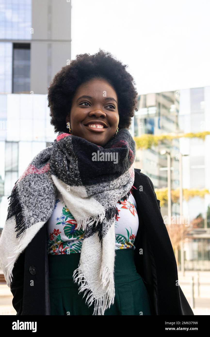 Allegra giovane afroamericana con acconciatura afro in abiti caldi e sciarpa in piedi sulla strada contro gli edifici moderni Foto Stock