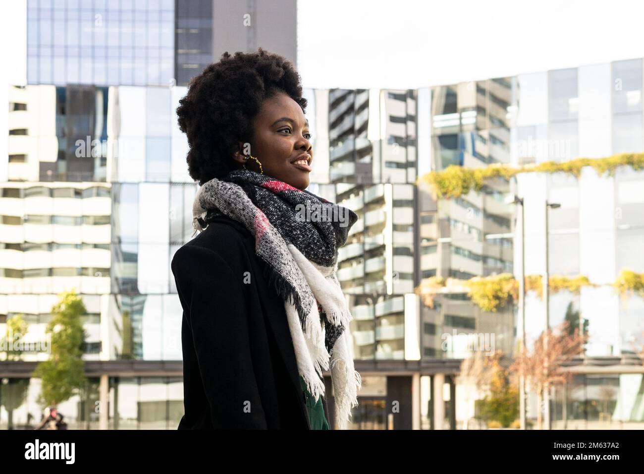 Vista laterale di allegra giovane afroamericana con acconciatura afro in abiti caldi e sciarpa in piedi sulla strada contro gli edifici moderni Foto Stock