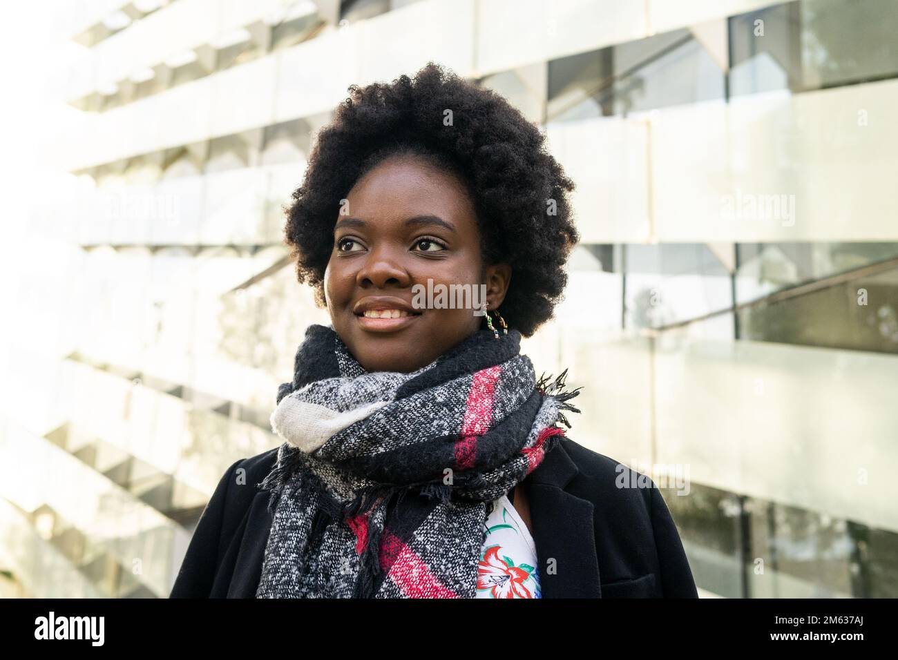 Allegra giovane afroamericana con acconciatura afro in abiti caldi e sciarpa in piedi sulla strada contro gli edifici moderni Foto Stock