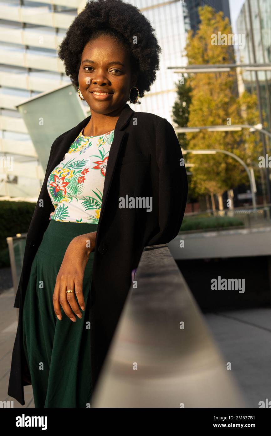 Positiva giovane afro-americana donna in giacca nera appoggiata su recinzione vicino a edificio moderno e guardando la macchina fotografica nelle giornate di sole Foto Stock