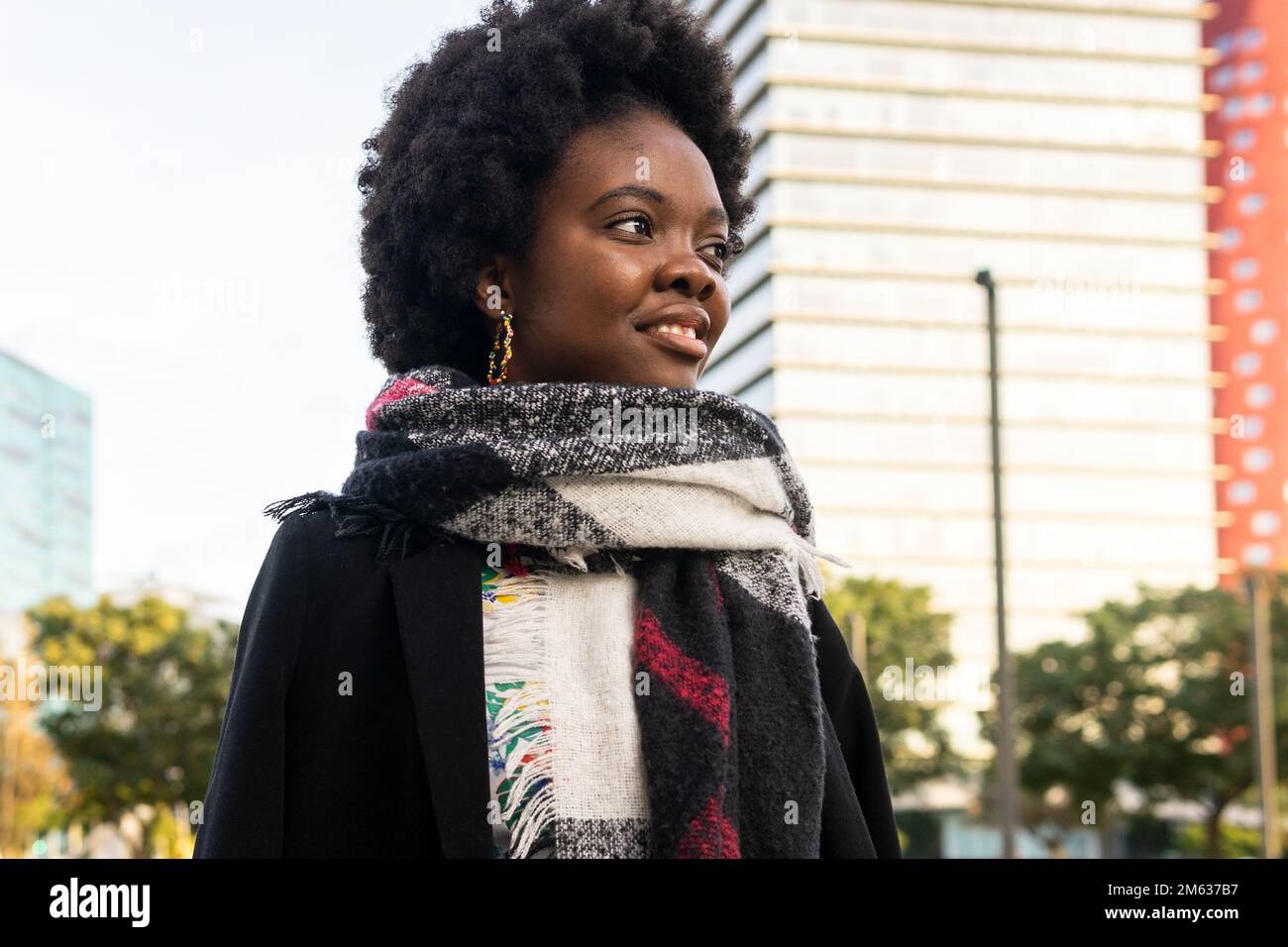Allegra giovane afroamericana con acconciatura afro in abiti caldi e sciarpa in piedi sulla strada contro gli edifici moderni Foto Stock