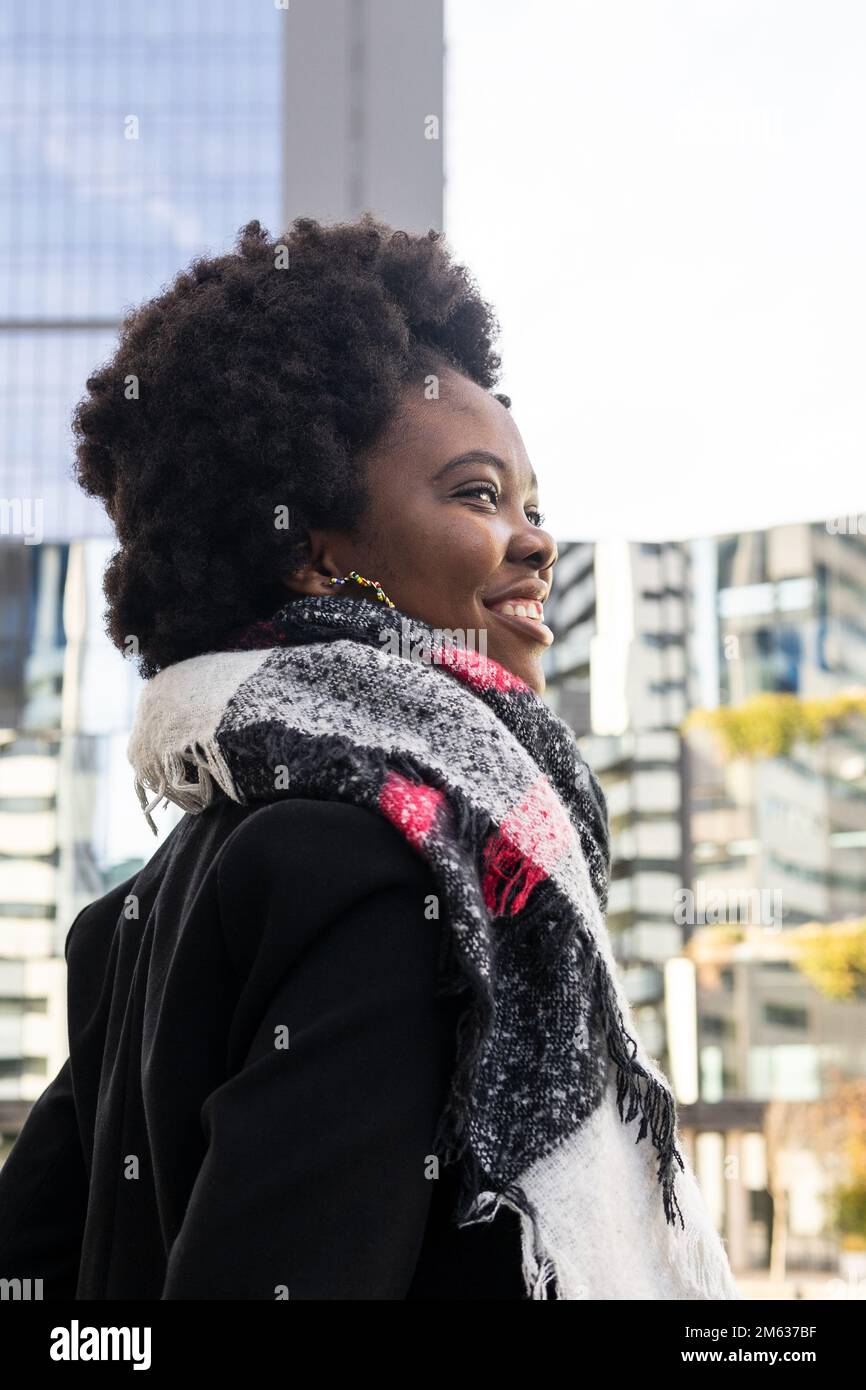 Vista laterale di allegra giovane afroamericana con acconciatura afro in abiti caldi e sciarpa in piedi sulla strada contro gli edifici moderni Foto Stock