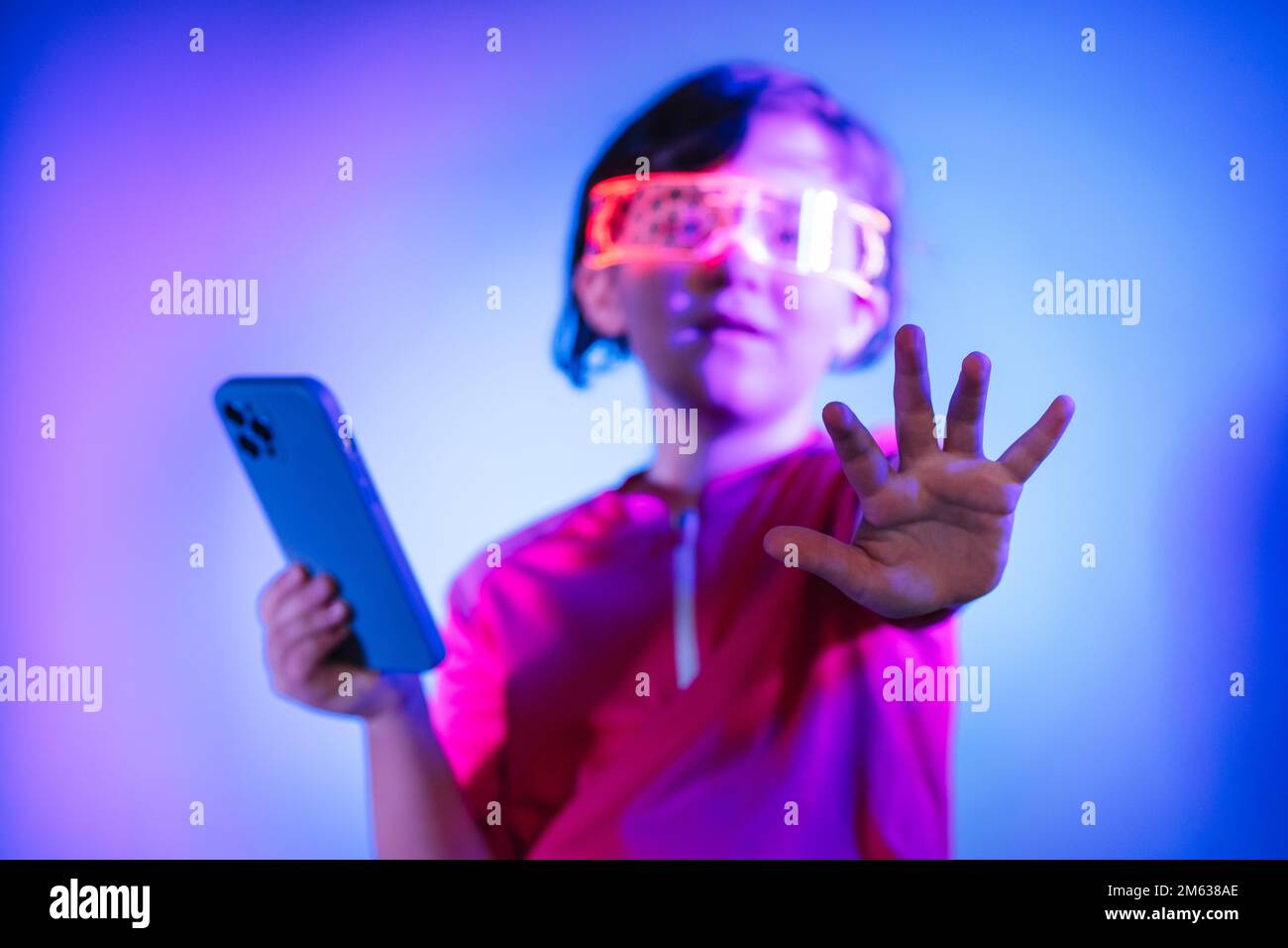Bambino con cellulare in occhiali intelligenti che gioca a videogame nel cyberspazio e tocca l'aria mentre si trova in studio con luci al neon blu Foto Stock