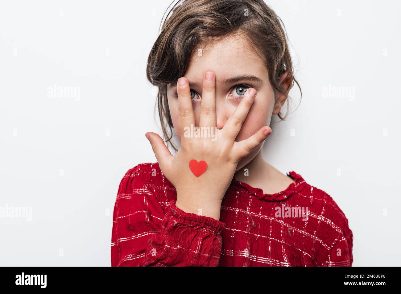 Adorabile ragazza con piccoli cuori sulle mani che coprono gli occhi mentre si gioca a nascondersi e cercare contro il muro bianco il giorno di San Valentino Foto Stock