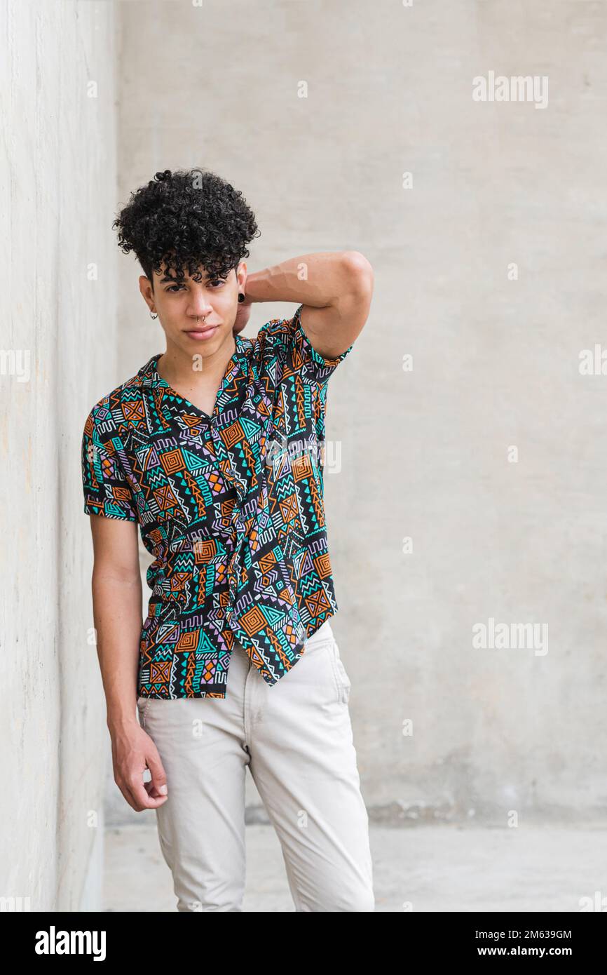 Uomo etnico in camicia e pantaloni con acconciatura afro in piedi contro parete di cemento guardando la macchina fotografica Foto Stock