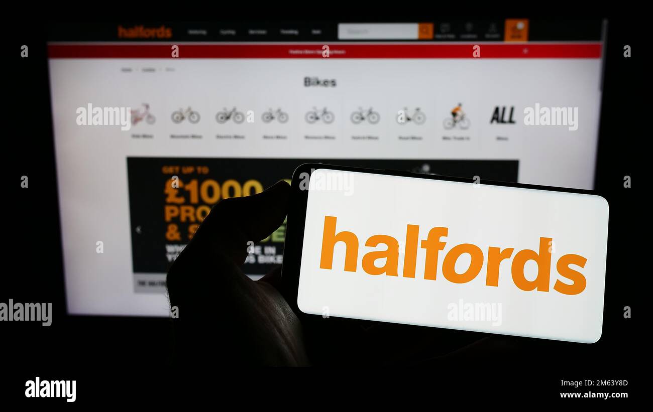 Persona che tiene il cellulare con il logo della società britannica di vendita al dettaglio Halfords Group plc sullo schermo di fronte alla pagina web aziendale. Messa a fuoco sul display del telefono. Foto Stock