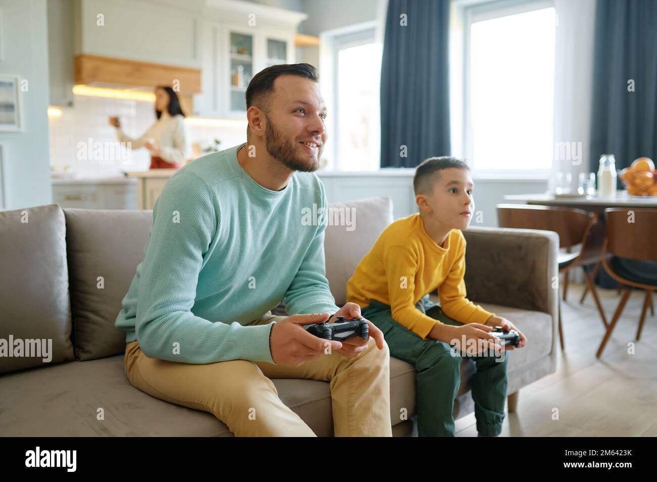 Papà e figlio che giocano a videogiochi mentre la mamma cucinava in cucina Foto Stock
