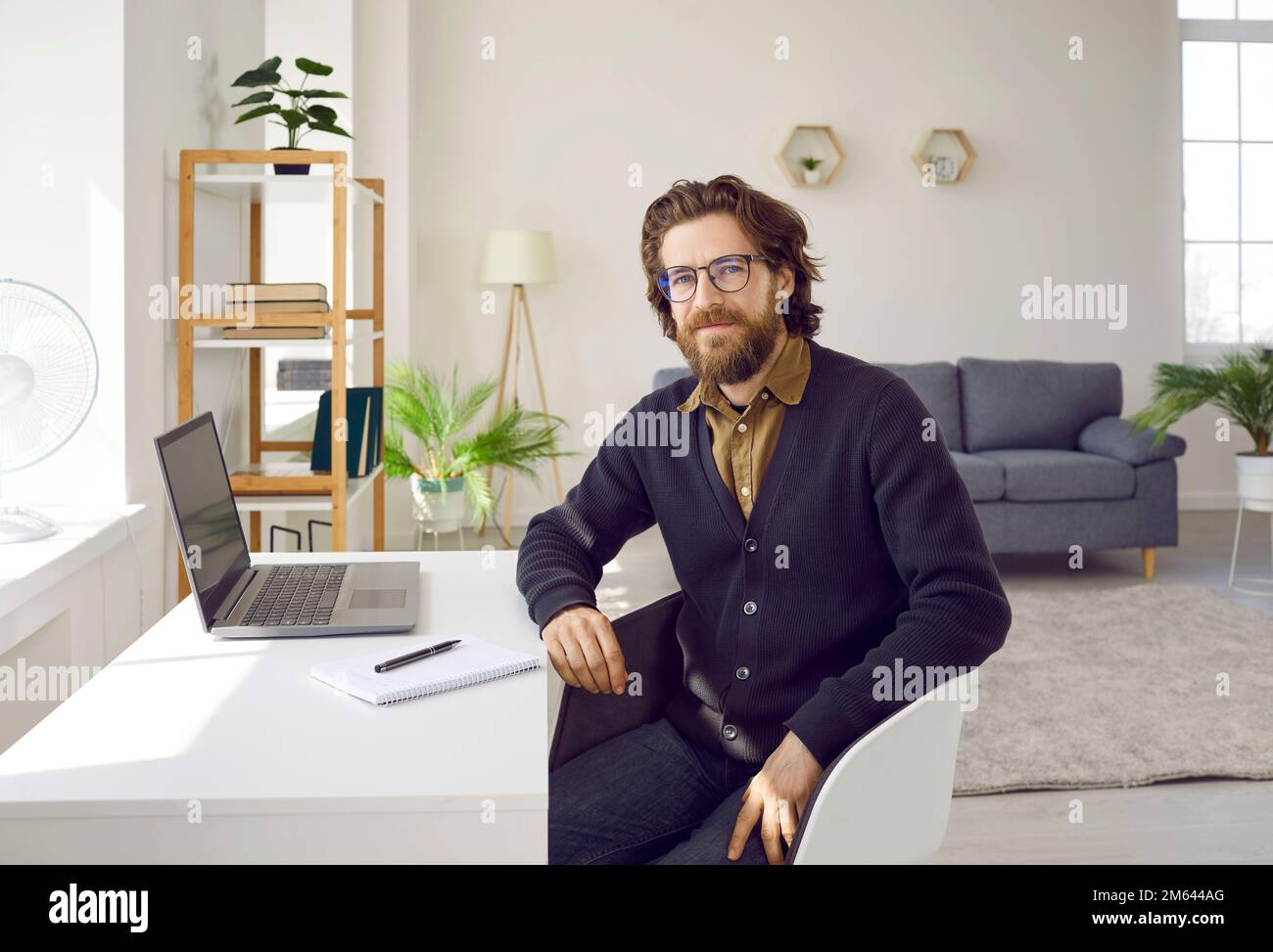 L'uomo di successo con barba e baffi guarda la fotocamera sulla scrivania con il computer portatile in ufficio Foto Stock