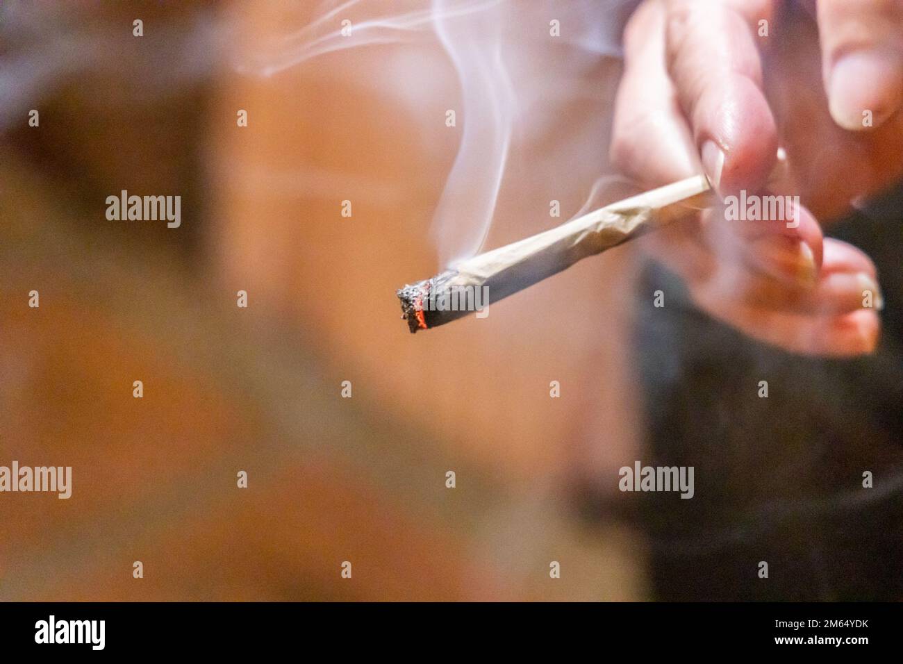Dito che tiene la sigaretta di cannabis illuminata con fumo in camera scura, con focalizzazione selettiva sulla cenere cannibica e il fuoco. Foto Stock