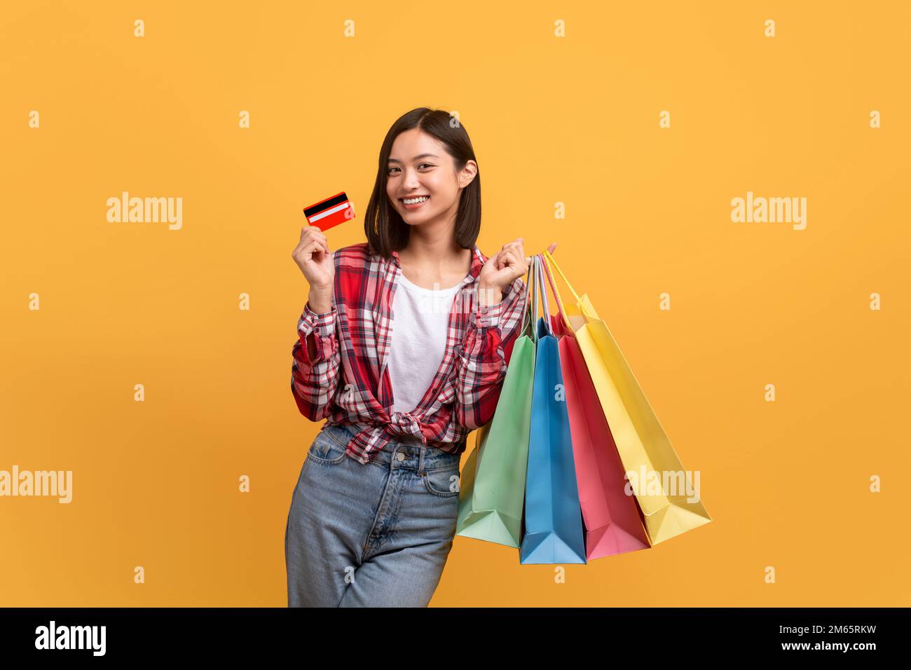 Concetto di rimborso. Donna asiatica felice posa con carta di credito e borse shopping colorate su sfondo giallo studio Foto Stock