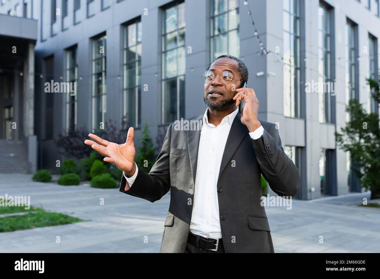 Allegro e soddisfatto boss afro-americano fuori ufficio edificio sorridendo e parlando con i colleghi al telefono, uomo in tuta d'affari a piedi durante la pausa pranzo. Foto Stock