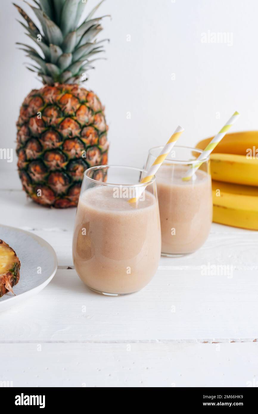 Due bicchieri di frullato di banana e ananas su un tavolo di legno bianco. Concetto minimo di dieta disintossicante, bevanda estiva di vitamina. Primo piano. Foto Stock