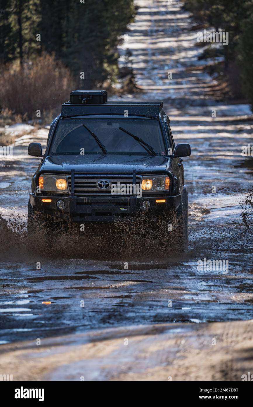 Vista frontale ad angolo del SUV fuoristrada Overland Land Cruiser che attraversa l'acqua su un sentiero innevato nella foresta Foto Stock