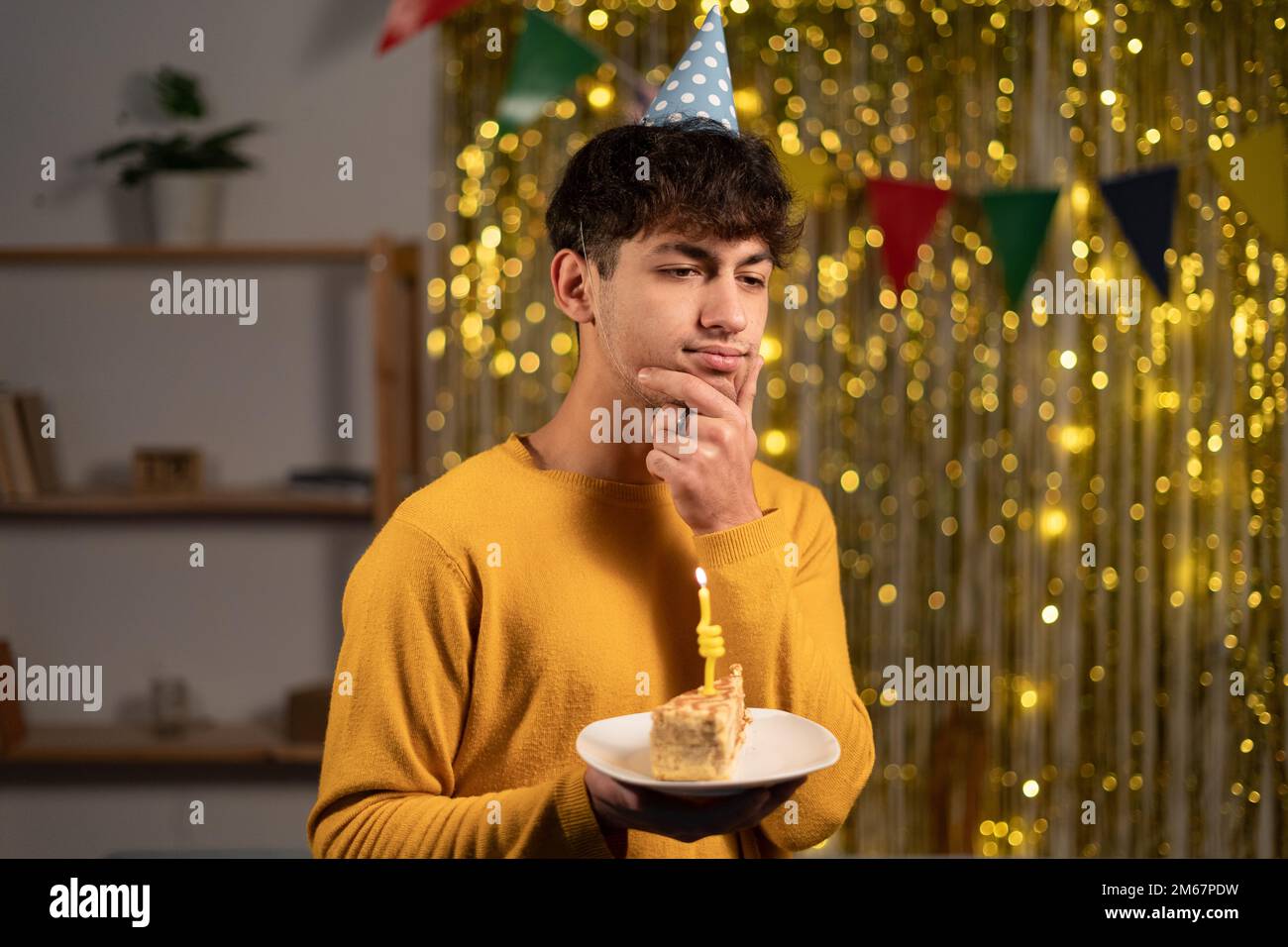 Giovane uomo premuroso che festeggia il suo compleanno tenendo un pezzo di torta che tocca il mento e il pensiero. Feste e feste Foto Stock