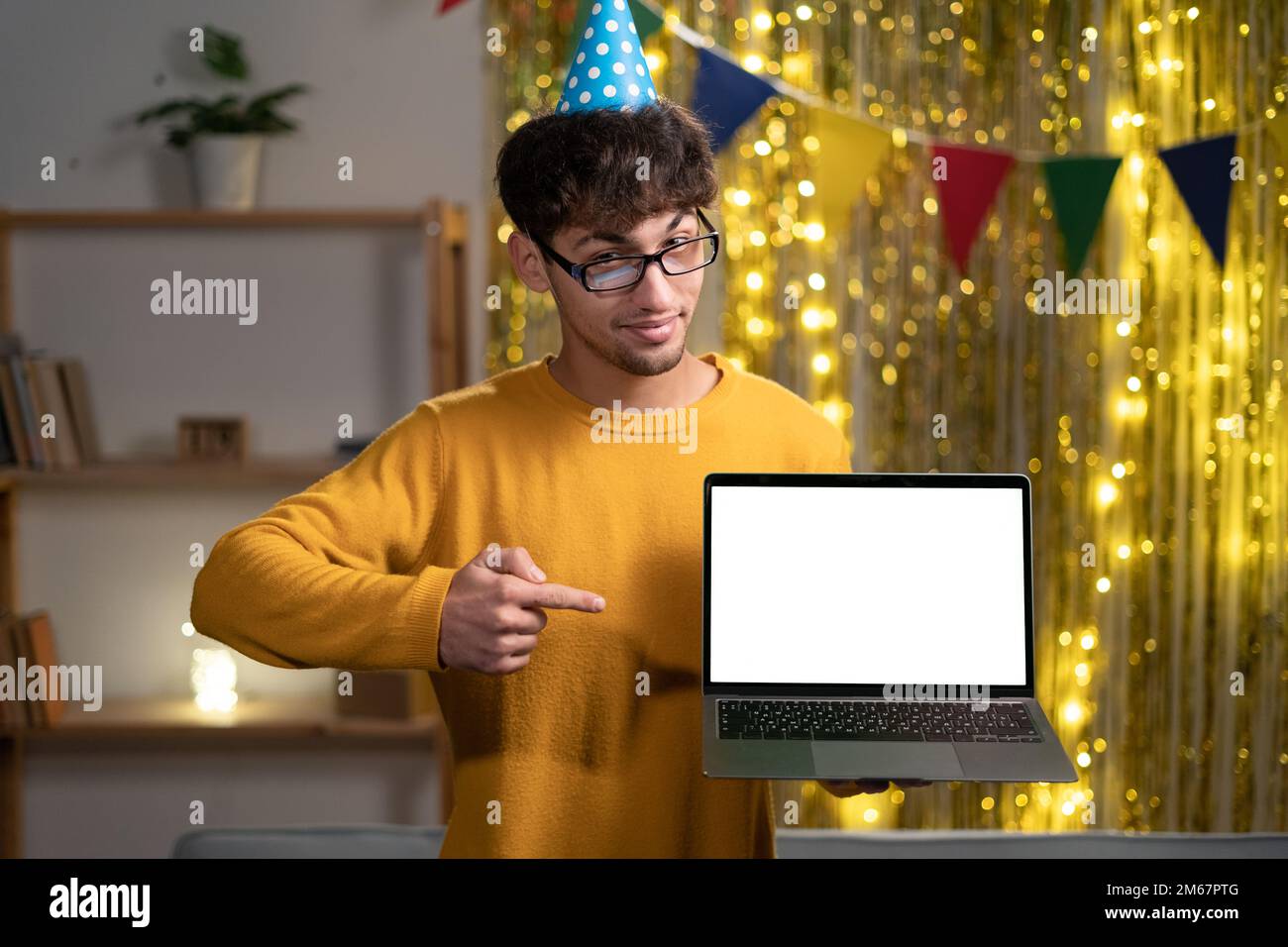 Ragazzo arabo o indiano gioioso in cappello da festa, freelance o studente, tenendo un computer portatile aperto con spazio vuoto mock-up e punta il dito su di esso, celebrare Foto Stock