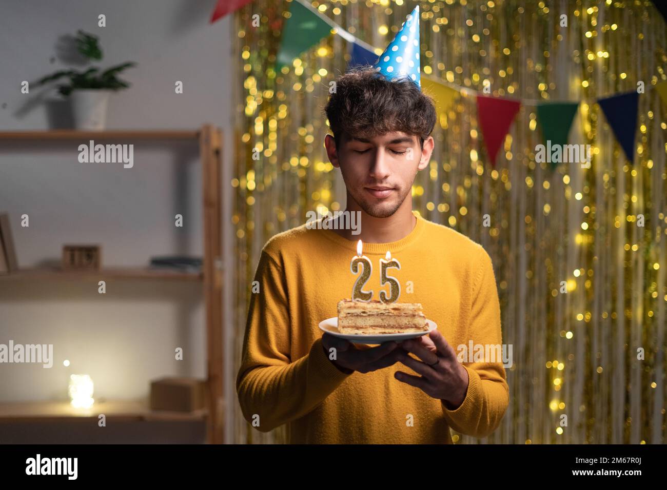 Ragazzo che festeggia 25st compleanno a casa, tenendo una torta con le candele e facendo un desiderio Foto Stock