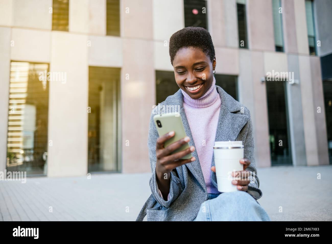 Donna afro-americana felice in outerwear elegante con tazza di caffè senza sprechi. Sorride e naviga sui social media sul cellulare mentre si siede su una panchina per strada in città al mattino Foto Stock