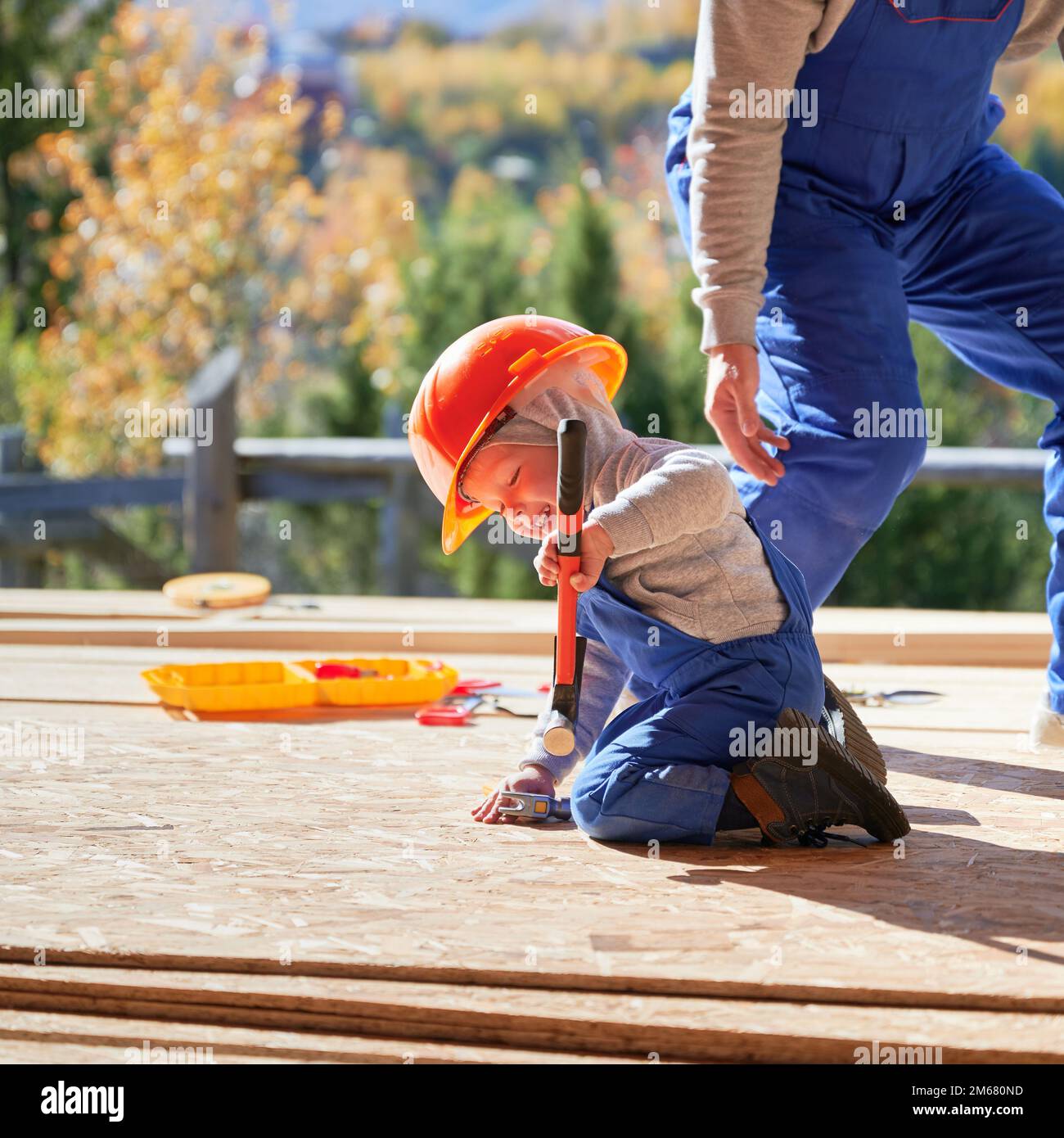 Padre con bambino figlio costruzione legno cornice casa. I costruttori maschi martellano il chiodo in asse sul cantiere, indossando casco e tute blu il giorno di sole. Carpenteria e concetto di famiglia. Foto Stock