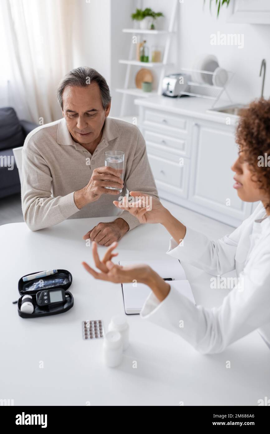curly afroamericano medico gesturing mentre parlando con il paziente di mezza età che tiene il vetro di acqua, immagine di scorta Foto Stock