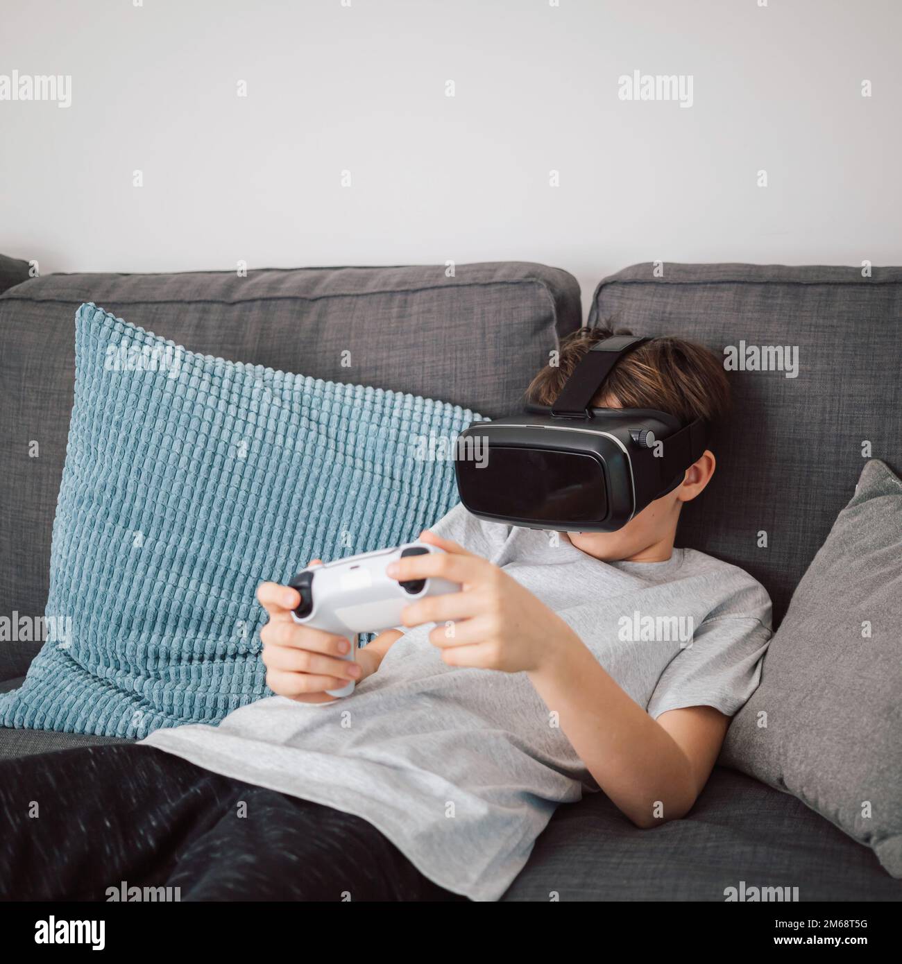 Bambino caucasico che indossa un visore VR e tiene un gamepad mentre si siede in salotto, giocando a giochi di simulazione di realtà virtuale Foto Stock