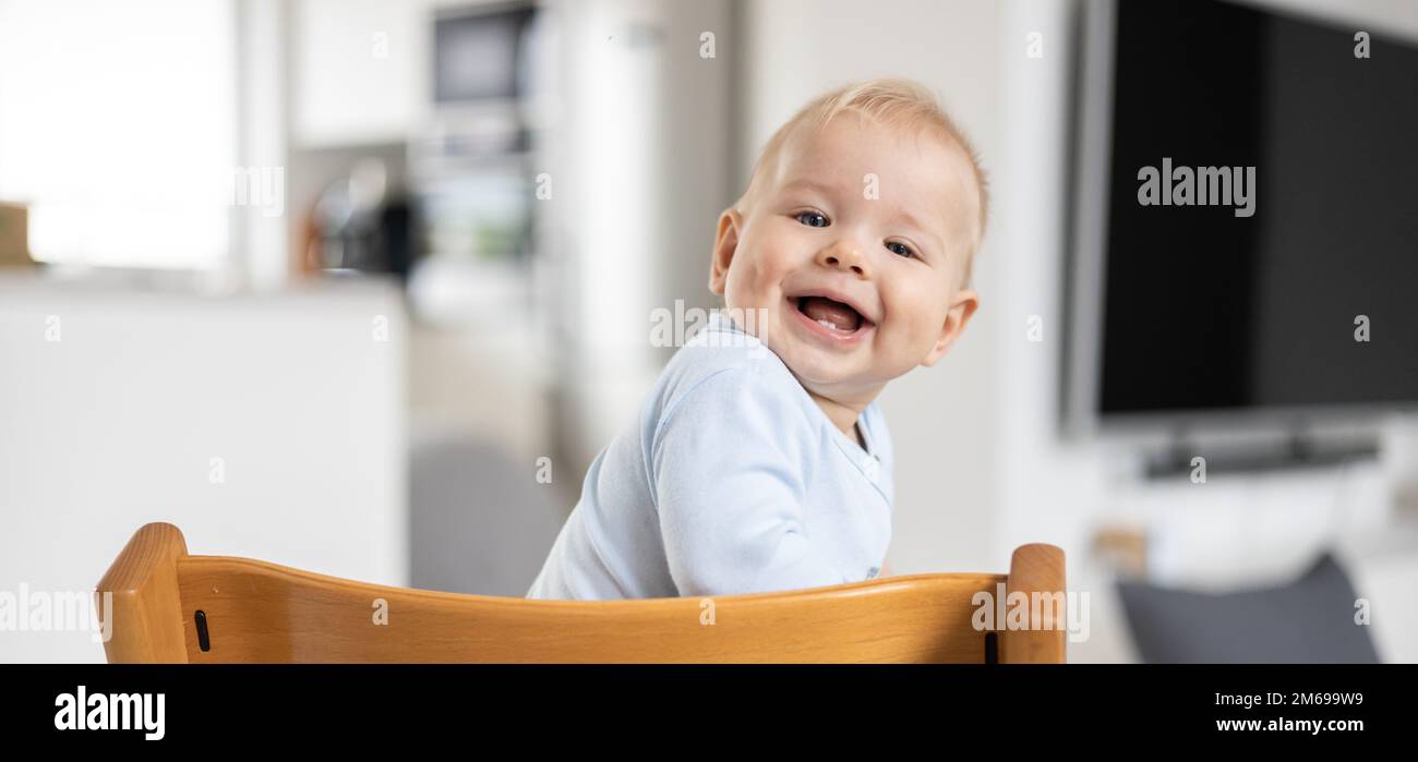 Felice bambino seduto in tradizionale design scandinavo seggiolone di legno e ridendo forte in una moderna casa luminosa. Sorriso carino del bambino Foto Stock