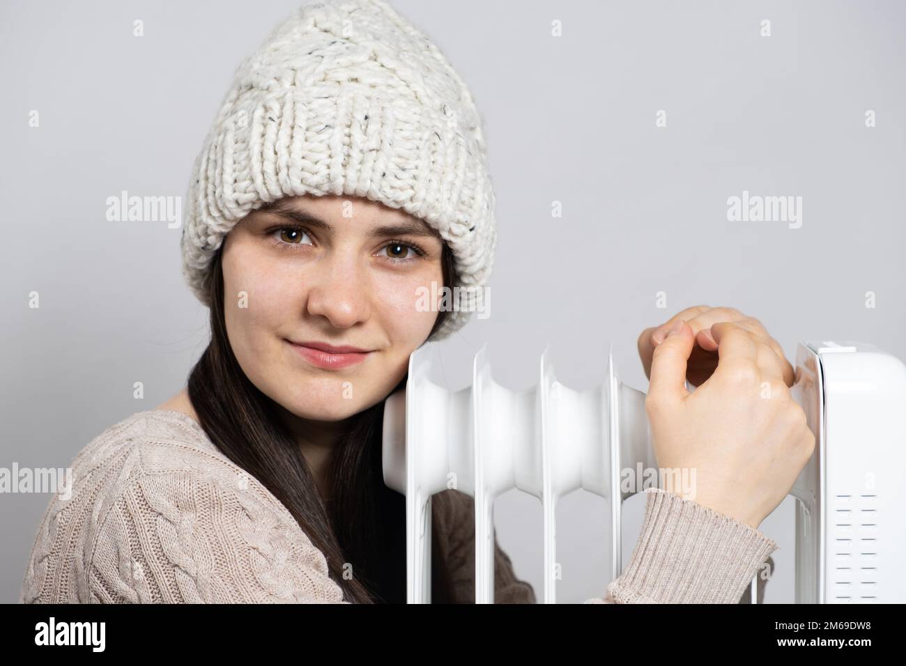 Una donna brunetta in un cappello abbraccia un riscaldatore ad olio, si riscalda in inverno. Foto Stock