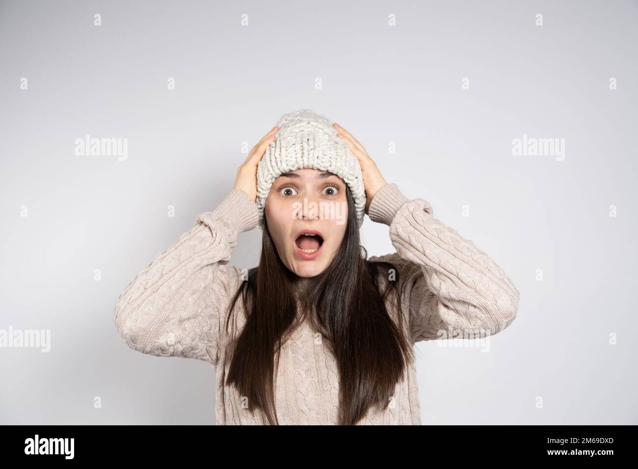 Una giovane bruna donna in un cappello e maglione guarda nella telecamera e urla tenendo le mani sulla testa. Foto Stock