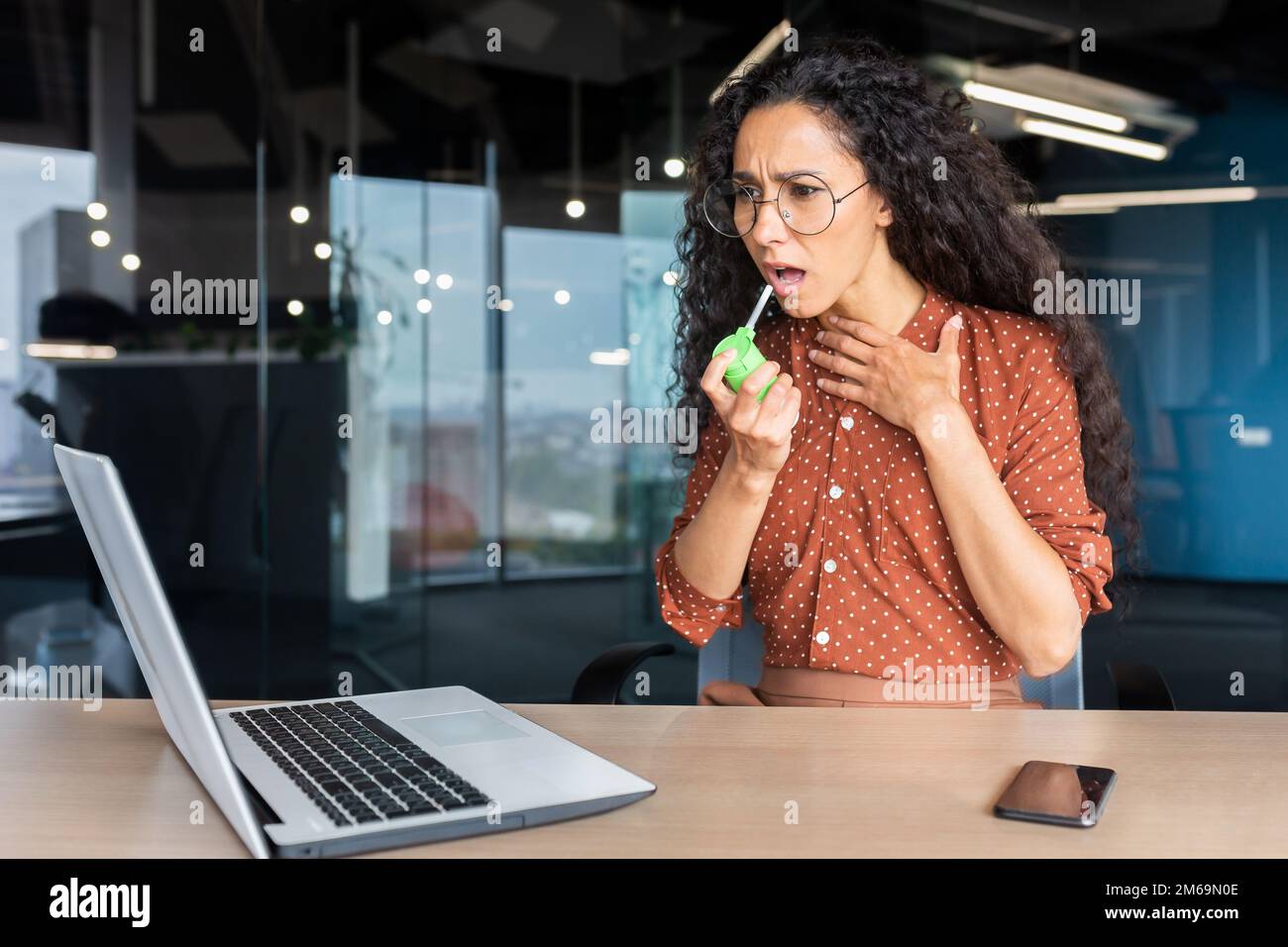 Donna ispanica ammalata in ufficio, donna d'affari ha mal di gola, mal di gola, usa la medicina per il sollievo dal dolore mentre si siede al lavoro in ufficio con un computer portatile al lavoro. Foto Stock