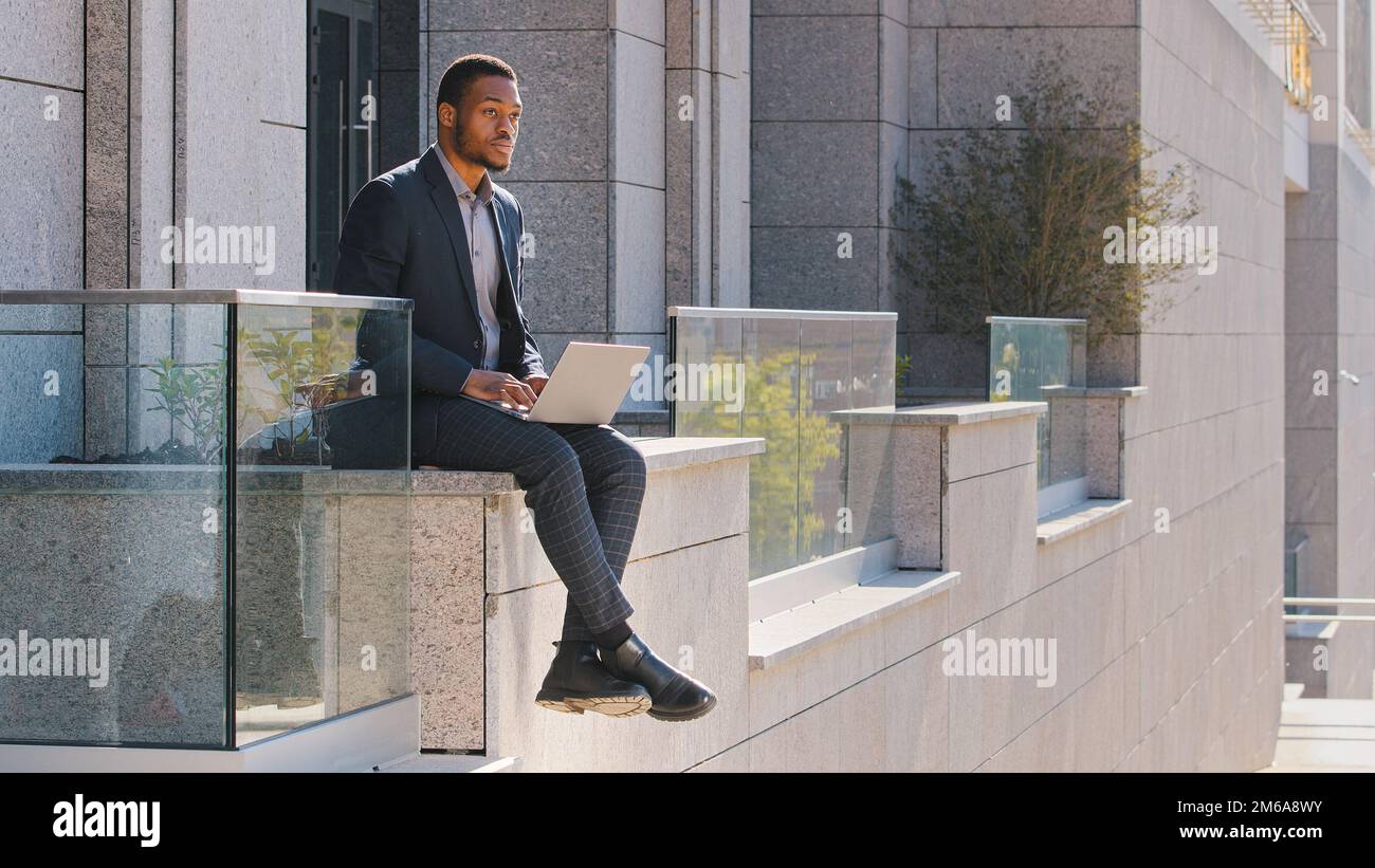Uomo d'affari etnico premuroso uomo d'affari afro-americano manager freelancer seduto all'aperto a digitare sul computer portatile preoccupato per il problema del tipo di lavoratore pensare Foto Stock
