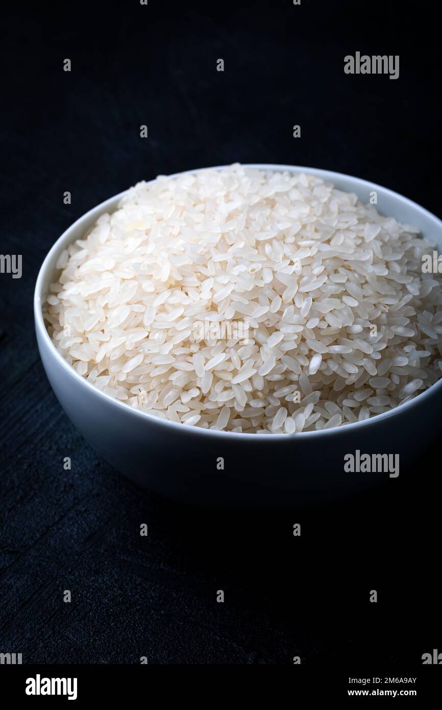 Recipiente bianco contenente riso bianco crudo su fondo nero. Foto Stock