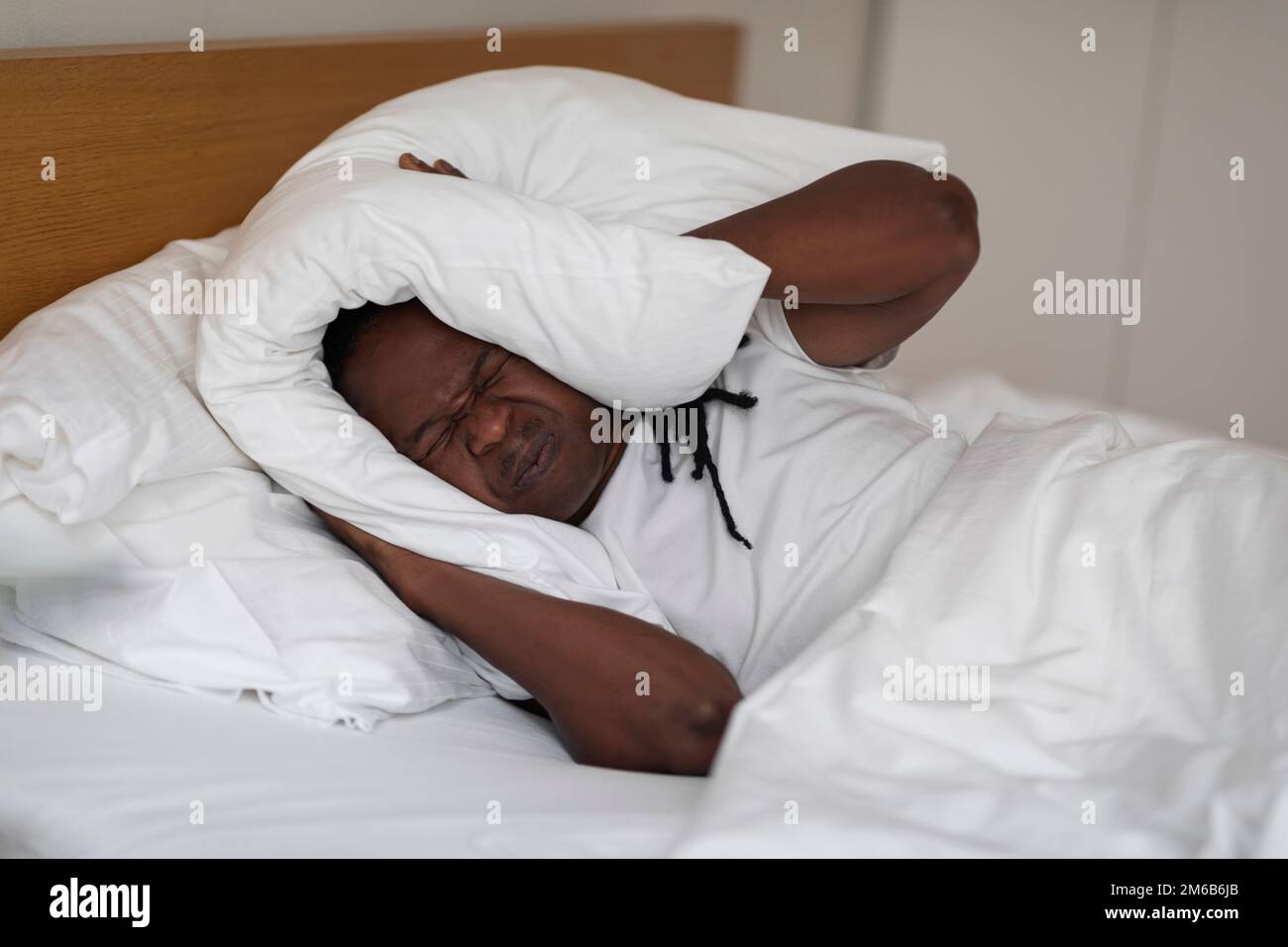 Irritato uomo nero sdraiato a letto e coprendo le orecchie con cuscino Foto Stock