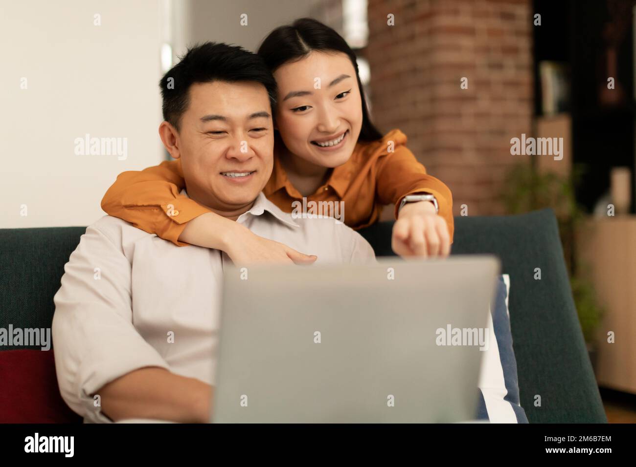 Sposi asiatici che amano fare acquisti su Internet, giovane donna che abbraccia il marito da dietro e indica lo schermo del notebook Foto Stock