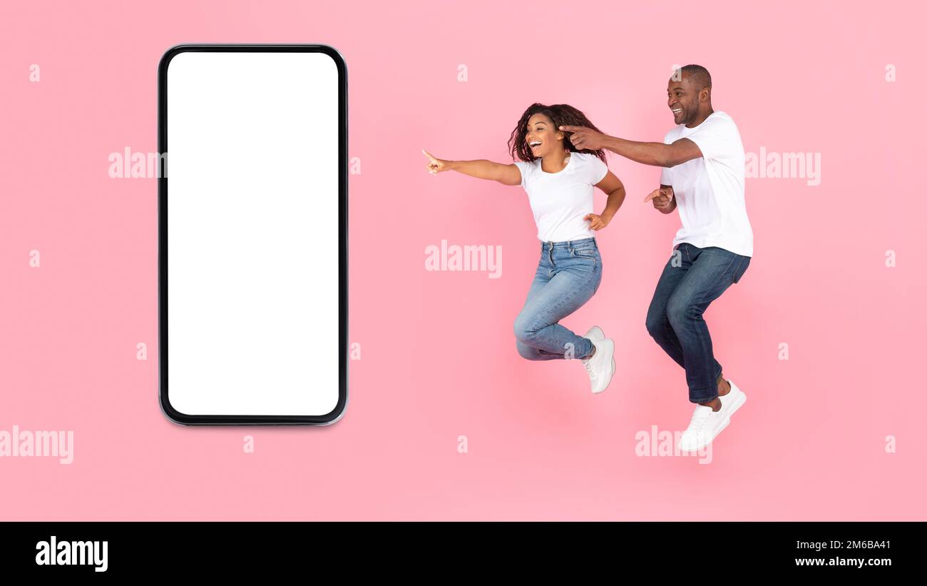 Overjoyed sposi afroamericani saltando in aria, puntando le dita al cellulare con schermo vuoto, mockup Foto Stock