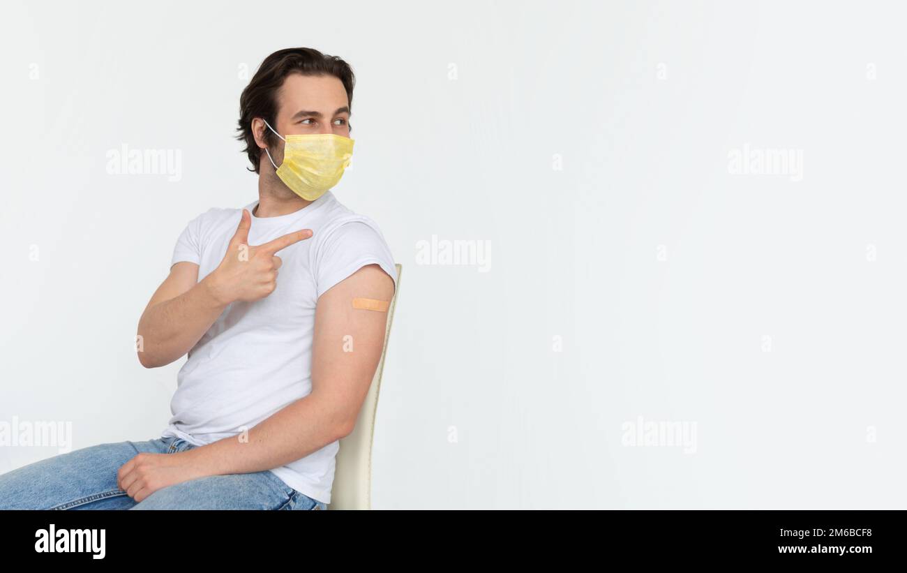 Felice giovane maschio caucasico in maschera protettiva con cerchietto sulla spalla dopo la vaccinazione mostra dito su spazio copia Foto Stock