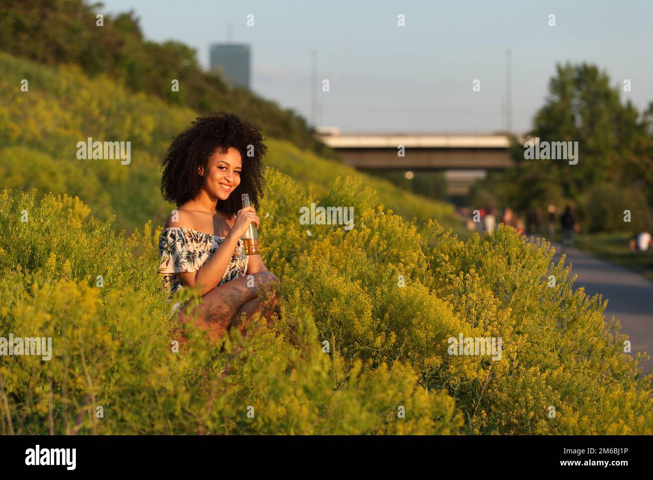 Bella ragazza africana con capelli ricci bere birra in un campo di fiori gialli Foto Stock