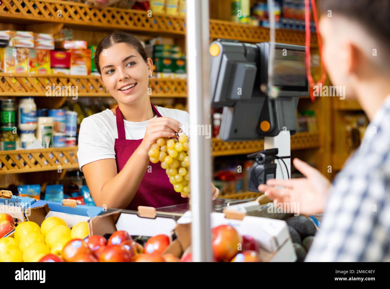 Donna venditore educato in piedi vicino bilance dietro il banco e pesando mazzo di uva per acquirente in supermercato Foto Stock