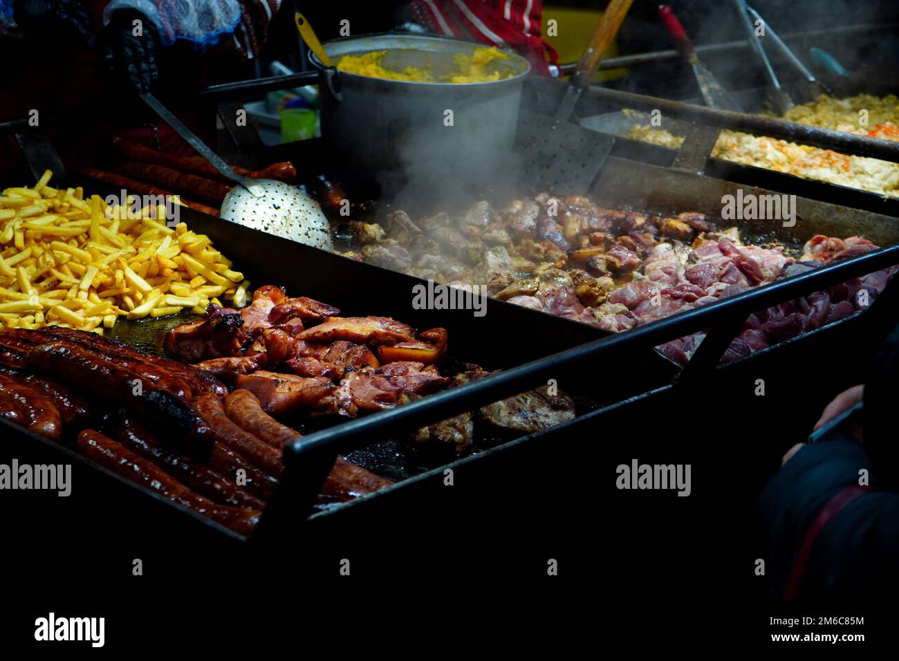 Cibo tradizionale rumeno che viene cucinato all'aperto per la vendita al mercato di Natale a Bucarest in serata. Carne di maiale, salsicce, polenta e patatine fritte. Foto Stock