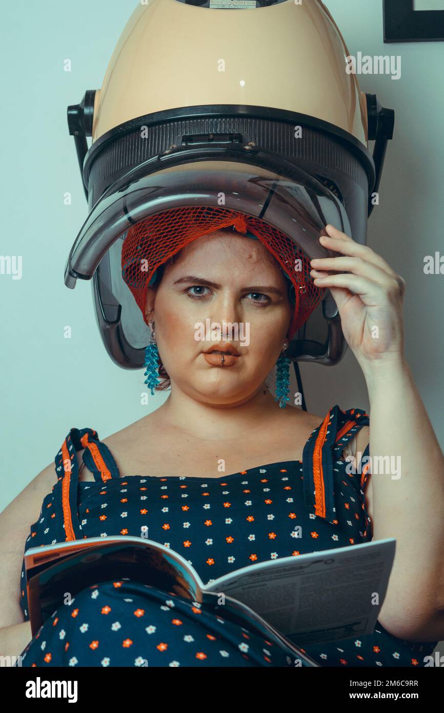 giovane donna d'epoca ispanica latina di etnia argentina, al parrucchiere facendo arricciacapelli su una macchina arricciacapelli, con una rivista in mano e lo Foto Stock