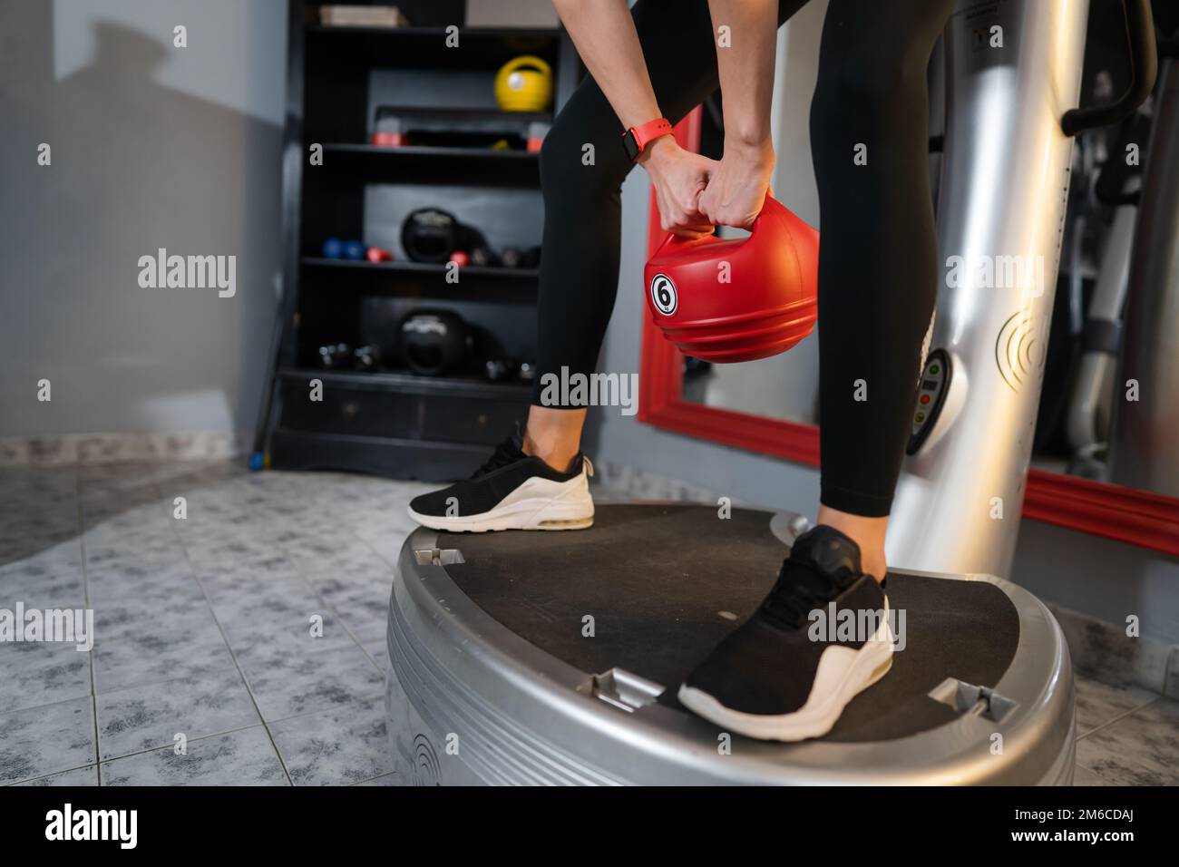 Donna sconosciuta tenere rosso Kettlebell peso mentre in piedi sulla piattaforma di vibrazione della piastra di potenza macchina durante l'allenamento squat esercizio copia spazio Foto Stock