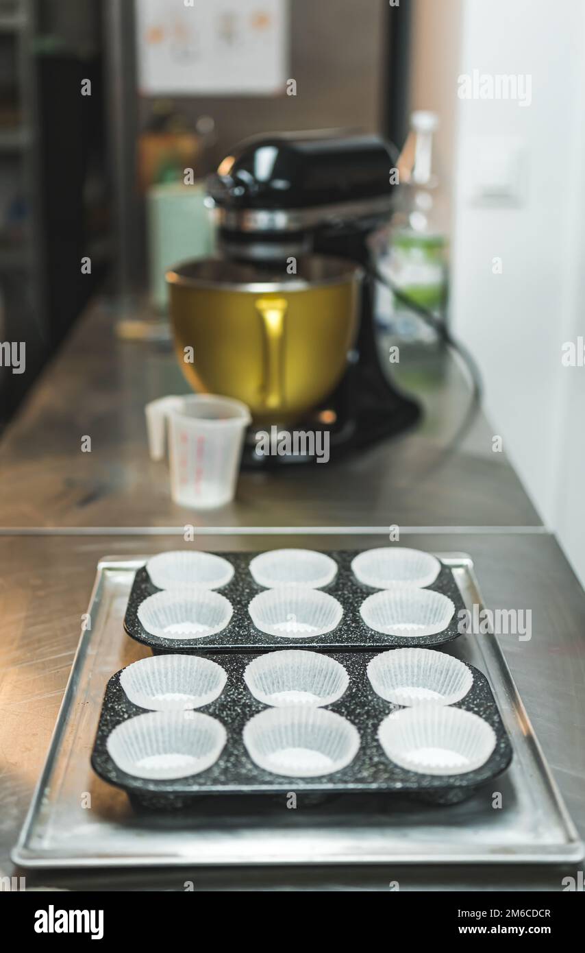 Fodere di cupcake preparate in una teglia su un vassoio da forno di metallo Mixer nero per alimenti e tazza di plastica sfocata sullo sfondo. Foto di alta qualità Foto Stock