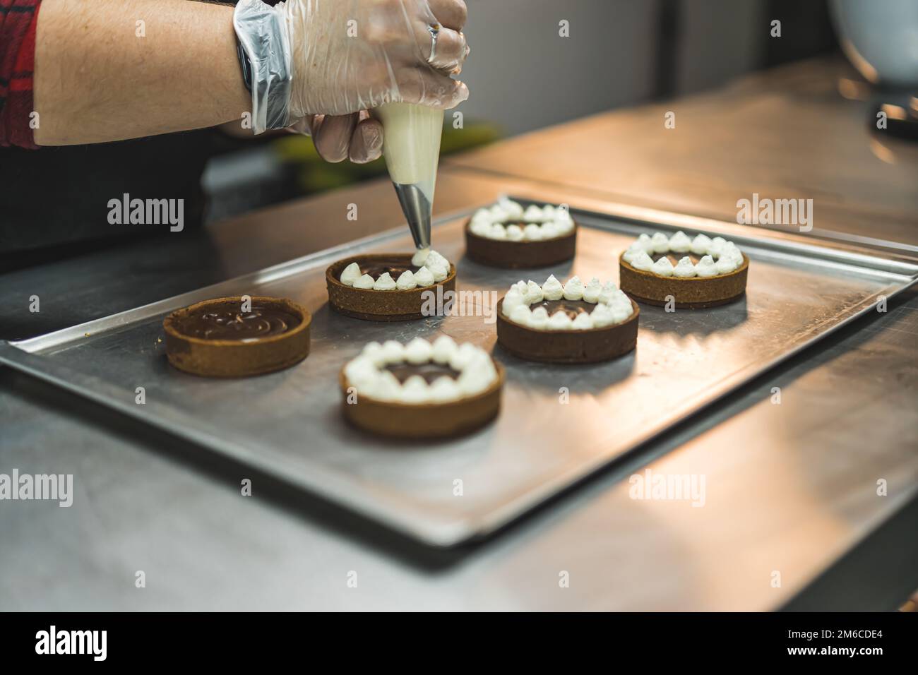 Un panettiere con guanto trasparente che decora le crostate poste su una teglia da forno con crema bianca utilizzando un sacchetto per pasticceria. Sfondo sfocato. Foto di alta qualità Foto Stock