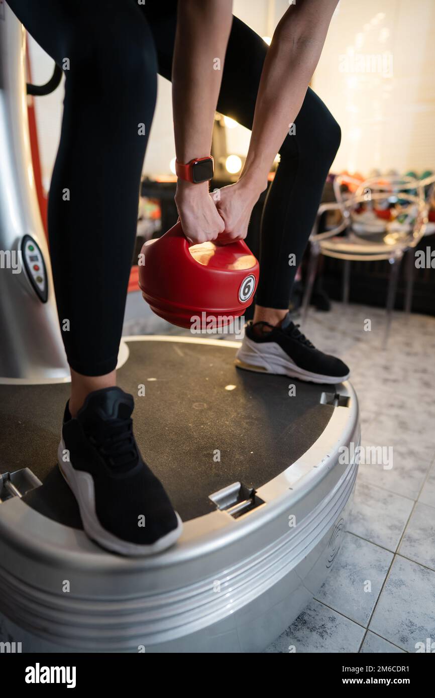 Donna sconosciuta tenere rosso Kettlebell peso mentre in piedi sulla piattaforma di vibrazione della piastra di potenza macchina durante l'allenamento squat esercizio copia spazio Foto Stock