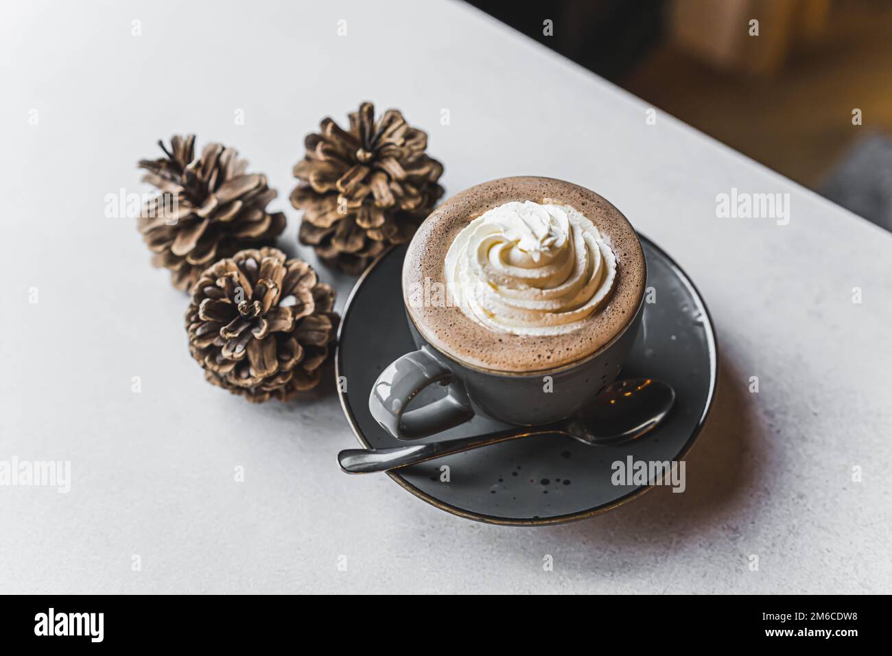Caffè decorato con panna montata in una tazza grigia su un piatto grigio con un cucchiaio. Tre coni posti accanto alla coppa. Foto di alta qualità Foto Stock
