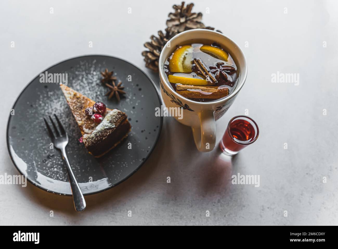 Dessert e una bevanda calda. Un pezzo di torta decorato con zucchero a velo e frutta posta su un piatto nero accanto ad una tazza bianca con una bevanda calda di vino e un piccolo bicchiere di sciroppo rosso. Foto di alta qualità Foto Stock